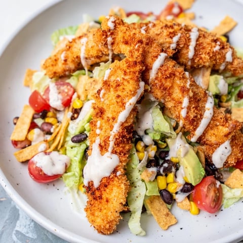 Crispy Chicken Taco Salad features golden-brown spiced chicken strips piled on fresh romaine lettuce. Topped with tortilla strips, cherry tomatoes, black beans, corn, cheddar, avocado, and creamy salsa ranch dressing.
